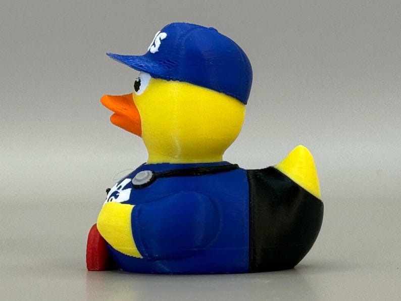 May include: A yellow rubber duck wearing a blue baseball cap with white lettering and a blue uniform. The duck has an orange beak and red feet. The duck is a novelty item.