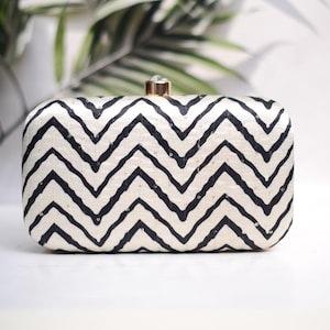 Puede incluir: Un clutch blanco y negro con un patrón de chevron. El clutch tiene un cierre dorado con un strass transparente.