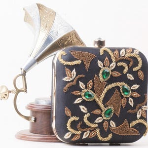 Peut inclure: Une pochette en satin noir avec des broderies dorées et vertes. La pochette est de forme carrée et se ferme à l'aide d'un fermoir. La broderie représente un motif floral avec des feuilles et des fleurs.