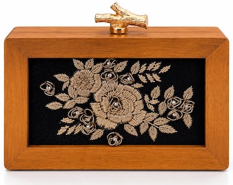 Könnte beinhalten: Eine rechteckige Clutch aus Holz mit einem glatten hellbraunen Rahmen und einem goldfarbenen Metallverschluss in Astform. Das Mittelteil besteht aus schwarzem Stoff mit aufwendigen Goldfadenstickereien in floralen Mustern.