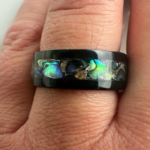 May include: A black ring with iridescent abalone shell inlay. The ring features a wide band with a glossy finish. The abalone shell displays shades of blue, green, and gold, creating a striking contrast against the black.