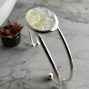 May include: A silver cuff bracelet with a large, oval-shaped centerpiece filled with a cluster of clear, iridescent stones. The bracelet has a sleek, modern design with smooth, curved lines and small ball accents.