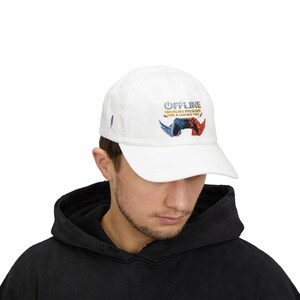 Puede incluir: Gorra de béisbol blanca con la palabra "OFFLINE" en azul y un gráfico de un mando con alas azules y rojas. El texto "ENJOY MY PRESENCE FOR A LIMITED TIME" también está en la gorra.