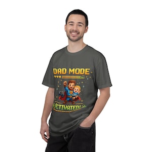 Può includere: T-shirt grigio scuro con un design pixelato. La maglietta presenta la scritta "DAD MODE ACTIVATED!" sopra un'illustrazione di un padre e un bambino supereroi. Il padre tiene in mano un controller di gioco e il bambino sorride.