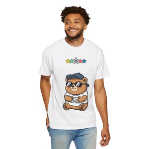 May include: White t-shirt featuring a cartoon bear wearing sunglasses and a cap, holding a game controller. The bear is brown with a white shirt. Above the bear are colorful stars. The shirt is worn by a person.