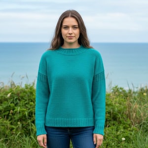 Könnte beinhalten: Ein türkisfarbener Pullover mit Rundhalsausschnitt und langen Ärmeln. Der Pullover hat gerippte Details an den Manschetten und am Saum. Die Person trägt eine blaue Jeans. Der Hintergrund zeigt ein Gewässer und grünes Laub.