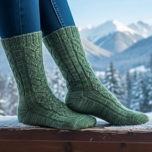 Puede incluir: Un par de calcetines verdes de punto trenzado. Los calcetines se llevan con vaqueros azules. Tienen un patrón texturizado y llegan hasta la rodilla. El fondo muestra un paisaje nevado con montañas y árboles.