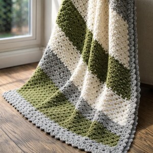May include: A handmade crochet blanket with a geometric pattern. The blanket features alternating stripes of olive green, cream, and gray. The edges are finished with a scalloped border. The blanket is draped over a wooden surface.