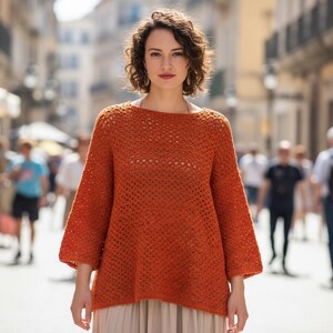 May include: A woman in a rust-coloured, open-weave crochet sweater with wide sleeves and a boat neck. The loose-fitting sweater is the focal point. The background is a blurred street scene with people and buildings.