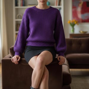 May include: A ribbed, purple sweater with long sleeves and a crew neck is paired with a black mini skirt. The person is seated on a brown sofa in a well-lit room. The sweater's texture and color are prominent.