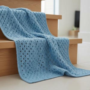 May include: A light blue crocheted blanket draped over wooden stairs. The blanket has a textured pattern with open spaces, creating a soft and inviting look. The blanket's color is a gentle, muted blue, suitable for home decor.