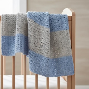 May include: A blue and gray knitted baby blanket draped over a wooden crib. The blanket features alternating stripes of light blue and gray, with a textured pattern. The crib is made of light-colored wood.