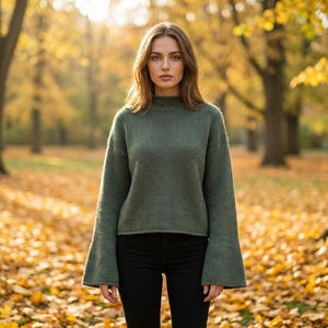 May include: A woman wearing a green sweater with flared sleeves and black pants stands in a park. The sweater has a high neckline. The background features trees with yellow leaves and fallen leaves on the ground.