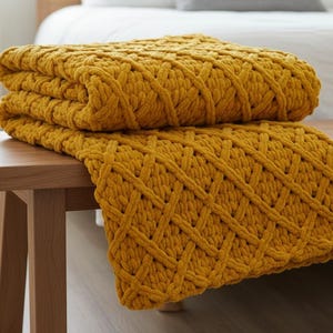 May include: A folded mustard yellow knitted blanket with a diamond pattern rests on a wooden bench. The thick, textured throw blanket is made of a soft material and is folded neatly. The blanket is a home decor item.