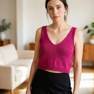 May include: A magenta-colored, knitted tank top with a V-neck. The top is sleeveless and has a ribbed hem. The person in the image is wearing a black skirt. The background includes a white couch, wooden furniture, and plants.