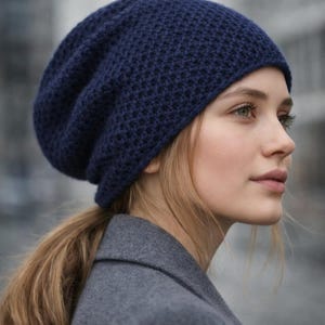 Op de afbeelding: Een marineblauwe gebreide beanie. De hoed heeft een textuurpatroon en wordt gedragen door een persoon met blond haar en een grijze jas. De hoed lijkt gemaakt van een zacht, warm materiaal, geschikt voor koud weer.