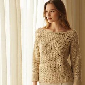 May include: A beige, long-sleeved sweater with a boat neck. The sweater has a textured, open-knit design. The sleeves have ribbed cuffs. The model is wearing the sweater in front of a sheer curtain.