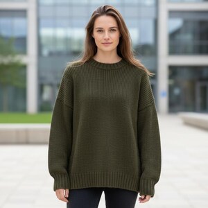 May include: An olive green sweater with a crew neck and long sleeves. The sweater has a ribbed hem and cuffs. The model is wearing black pants. The background is a modern building.