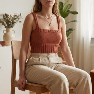 May include: A rust-colored, knitted crop top with wide straps and a square neckline. The top is paired with beige linen pants. The image is of a person sitting on a wooden chair in a well-lit room with plants.