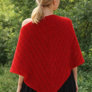 May include: A vibrant red knitted poncho with a textured pattern. The poncho is draped over the shoulders, showcasing its triangular shape and cozy design. The garment is made of a soft, warm material, perfect for cooler weather.
