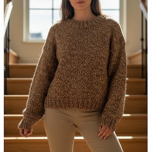 Puede incluir: Un suéter de punto grueso marrón con cuello redondo. El suéter tiene una apariencia texturizada y mangas largas. La modelo lleva pantalones color beige. El fondo incluye una escalera y una ventana.