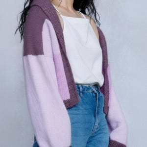 May include: A color-blocked cardigan in shades of purple and lavender. The hooded sweater features a solid dark purple hood and cuffs, with a lighter purple on the sleeves and a lavender body. The cardigan is worn over a white tank top and blue jeans.