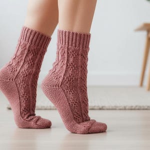 Op de afbeelding: Een paar stoffig roze gebreide sokken met een textuur van een ruitpatroon. De sokken worden gedragen op de voeten van een persoon, met de tenen en hielen zichtbaar. De sokken hebben een geribbelde boord en zijn gemaakt van zacht materiaal.