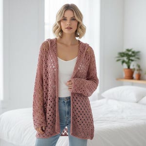 Puede incluir: Cárdigan con capucha de ganchillo rosa polvoriento. El diseño de tejido abierto presenta una longitud y mangas largas. El cárdigan se usa sobre una camiseta blanca y unos vaqueros azules. El fondo incluye una cama y una ventana.