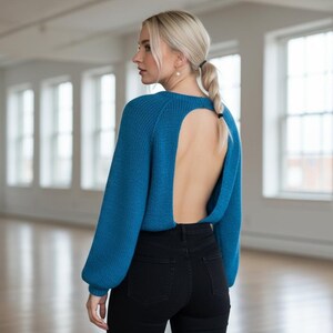 Könnte beinhalten: Ein tealblauer Pullover mit einem dramatischen offenen Rückendesign. Der Pullover hat lange Ärmel und wird mit schwarzen Jeans kombiniert. Das Model steht in einem hellen, offenen Raum.