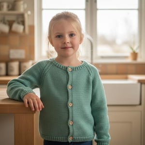 May include: A young child wearing a light green cardigan with wooden buttons. The cardigan is buttoned up and has a round neckline. The child is standing in a kitchen with a wooden countertop.