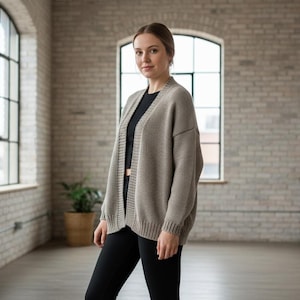 May include: A light brown open-front cardigan. The cardigan is worn over a black top and paired with black pants. The outfit is set against a backdrop of a brick wall and large windows, creating a casual, yet stylish look.