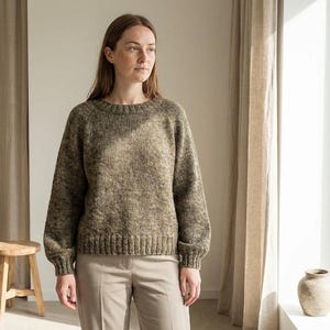 May include: A woman wearing a textured, olive green sweater with a crew neck and long sleeves. The sweater is paired with light-colored pants. A small wooden stool is in the background.