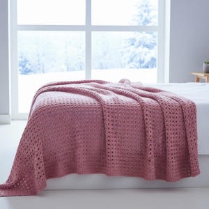 May include: A dusty rose-colored knitted blanket draped over a white bed. The blanket has a textured, open weave design. The bedroom has a large window with a snowy outdoor view, a white lamp, and a small wooden side table.