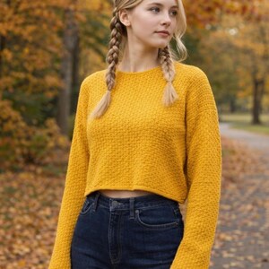May include: A mustard yellow long-sleeved cropped sweater with a textured knit pattern, paired with dark blue denim jeans. The sweater features a crew neckline and a straight hemline that sits above the waist, showcasing a simple, solid-colored design.