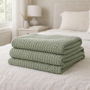 May include: Three sage green blankets with a woven texture, neatly stacked on a white bedspread. The image shows a bed, a bedside table, and a window in the background. Each blanket is folded.