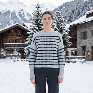 May include: A person wearing a light gray sweater with navy blue horizontal stripes and dark gray pants. The sweater has a crew neck and long sleeves. The background features snow-covered buildings and trees, with mountains in the distance.