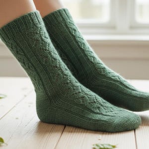 May include: A pair of green knit socks with a cable knit pattern. The socks are a solid, muted green color and appear to be made of a soft, warm material. The socks are on a light-colored wooden surface.