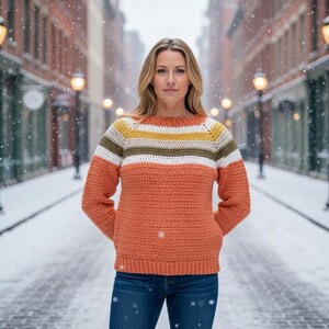 May include: A crochet sweater in shades of orange, white, yellow, and olive green, worn with blue jeans. The image shows a woman standing on a snow-covered street. The background features brick buildings and streetlights.