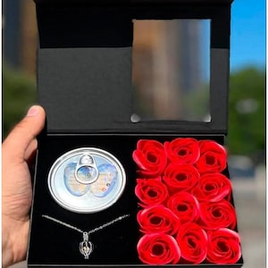 Op de afbeelding: Een zwarte cadeaudoos met een transparant venster aan de bovenkant. De doos bevat een zilveren blik met een deksel, een zilveren ketting met een parel hanger en een dozijn rode rozen.