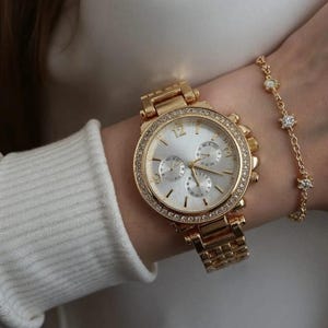 May include: Gold-toned wristwatch with a silver face and a bezel encrusted with small, clear crystals. The watch has a matching gold band and is paired with a delicate gold bracelet featuring small, clear, star-shaped accents.