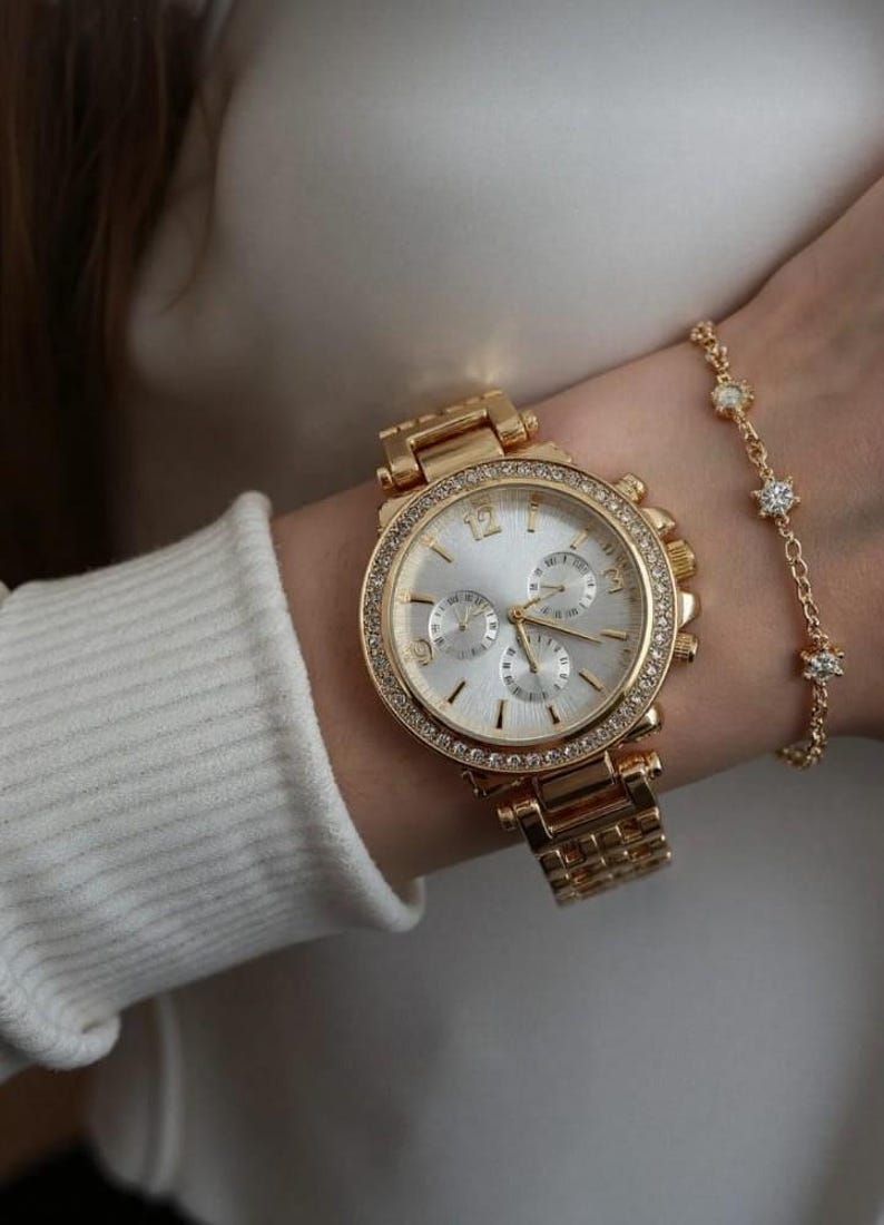 May include: Gold-toned wristwatch with a silver face and a bezel encrusted with small, clear crystals. The watch has a matching gold band and is paired with a delicate gold bracelet featuring small, clear, star-shaped accents.