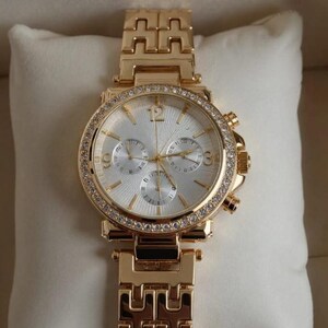 May include: Gold-toned wristwatch with a white face and a bezel encrusted with small, clear crystals. The watch has three sub-dials and a gold-toned link bracelet. The watch is displayed on a white pillow.