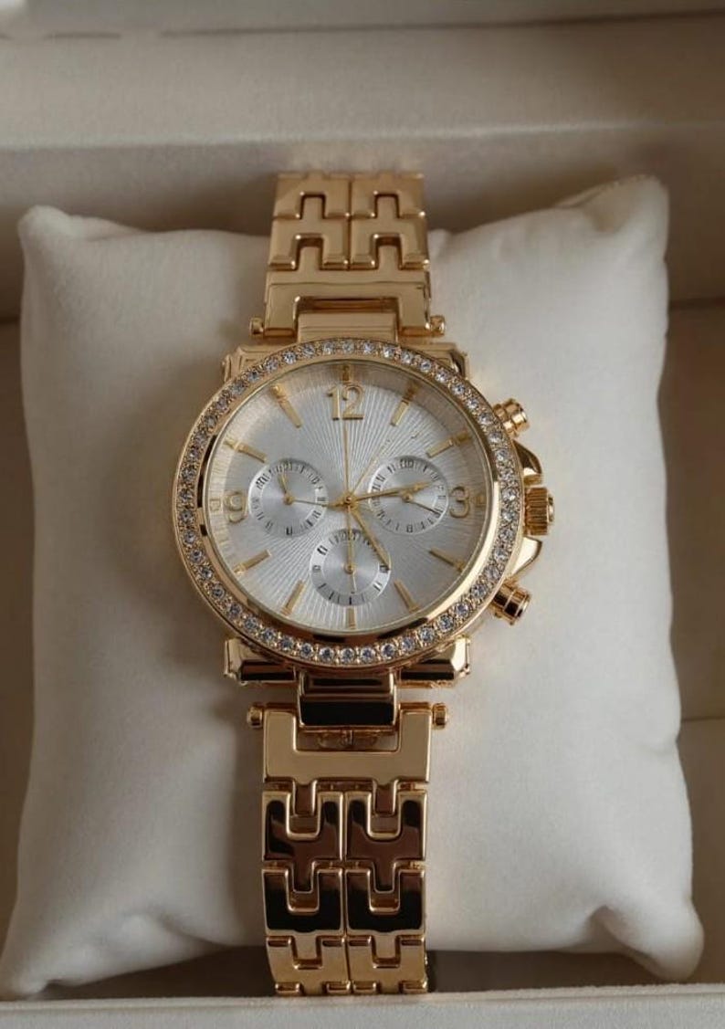 May include: Gold-toned wristwatch with a white face and a bezel encrusted with small, clear crystals. The watch has three sub-dials and a gold-toned link bracelet. The watch is displayed on a white pillow.