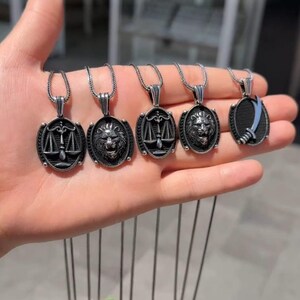 以下が含まれることがあります： さまざまなデザインのシルバーペンダントネックレス5本。ペンダントにはライオンの頭、天秤、剣が含まれています。ネックレスは手の上に表示されています。