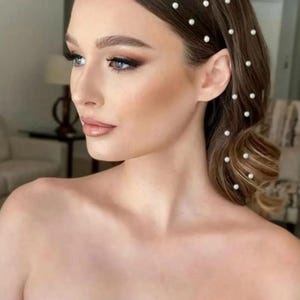 Peut inclure: Une femme aux cheveux bruns ornés de petites épingles à cheveux en perles blanches. Les cheveux sont coiffés avec une raie sur le côté et des ondulations douces. Elle a les yeux bleus et porte du maquillage. L'image est un gros plan de son visage et de ses épaules.