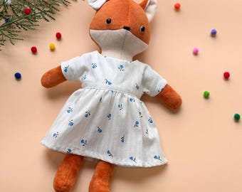 May include: A plush fox doll wearing a white dress with a blue floral pattern. The doll has a white bow on its head and is lying on a light orange surface.
