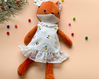 May include: A plush fox doll wearing a white dress with yellow flowers. The doll has a brown nose, black eyes, and a red bow on its head. The doll is lying on a light orange background.