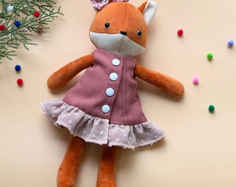 May include: A plush fox doll wearing a pink and white dress with white buttons. The doll has a red and white gingham bow on its head.