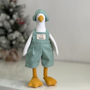 Peut inclure: Un jouet en peluche en forme d'oie portant un chapeau vert et une salopette. La salopette a une poche avec une étiquette blanche sur laquelle est écrit "Handmade".