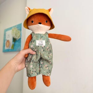 May include: A plush fox doll with orange fur, wearing a yellow hat and green floral overalls. The doll has a white muzzle and a small tag on the overalls. The doll is being held up by a person.